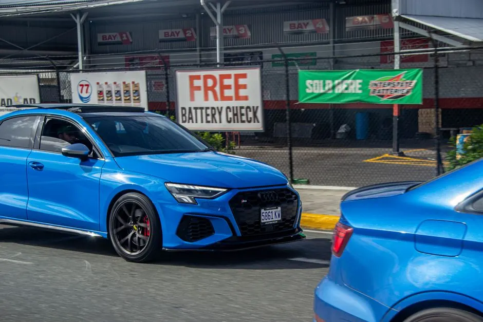 A vibrant blue Audi passes by an automotive service station offering free battery checks in Jamaica.